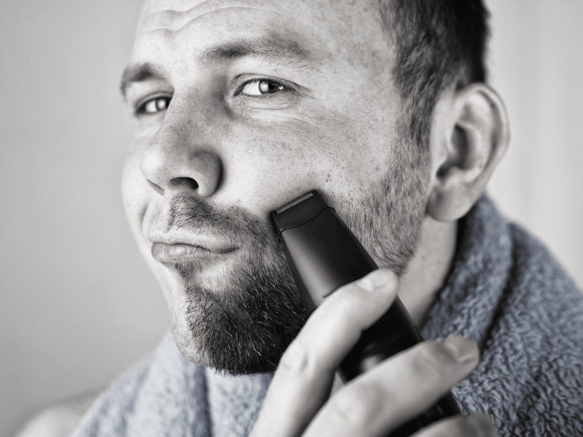 Man using beard trimmer on facial hair, grooming with towel around neck