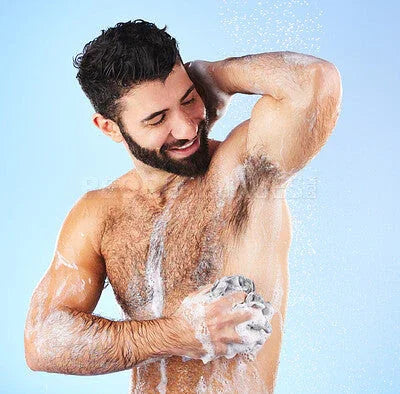 Smiling bearded man washing underarm with sponge and soap, foam and water splashing in shower