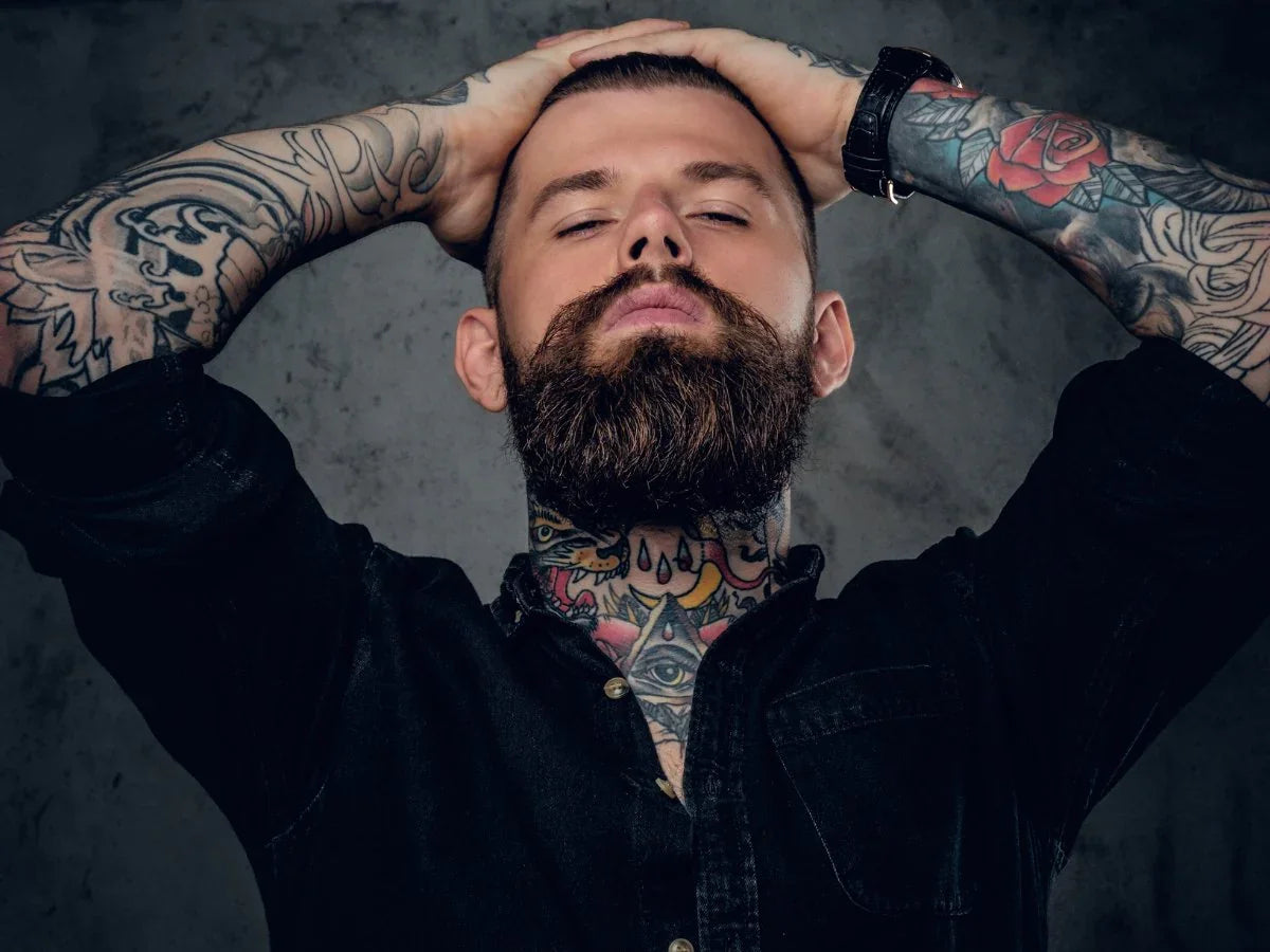 Tattooed man with a full beard and slicked-back hair posing against a dark background