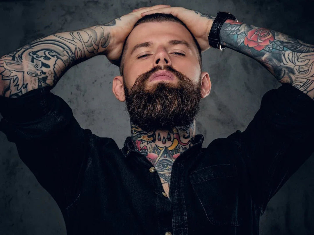 Tattooed bearded man in black shirt posing, showcasing groomed beard for Hairy Man Care.