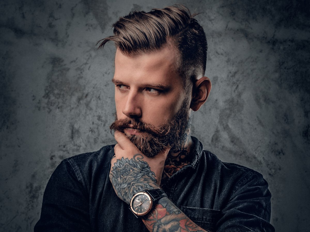 Stylish bearded man with tattoos and slicked back hair posing thoughtfully