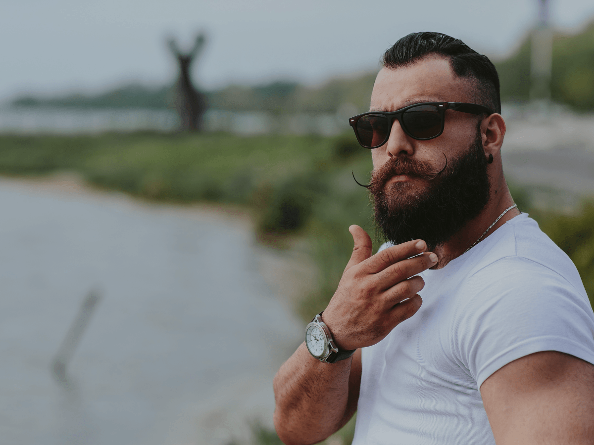 Man with thick, styled beard and mustache wearing sunglasses outdoors by water