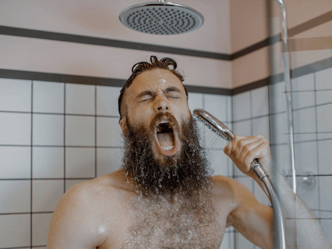 Bearded man washing beard in shower with handheld showerhead, tile background
