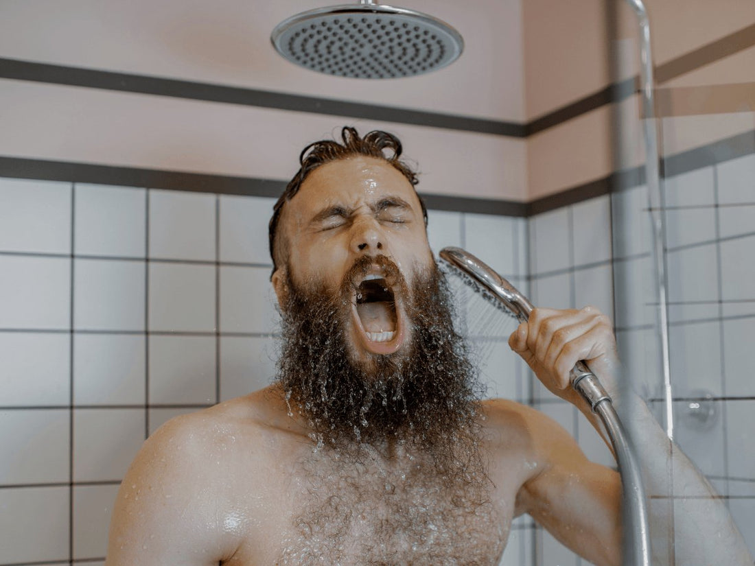 Bearded man washing beard in shower with handheld showerhead, tile background