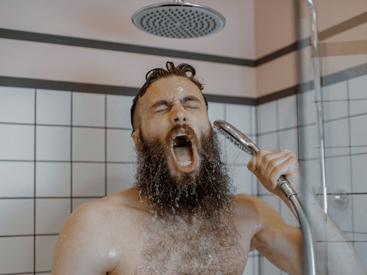 Bearded man washing beard in shower with handheld showerhead, tile background
