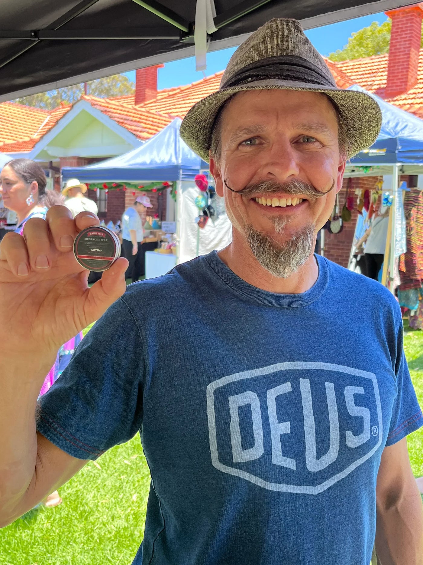 Smiling man with styled mustache holding Hairy Man Care moustache wax at outdoor market.