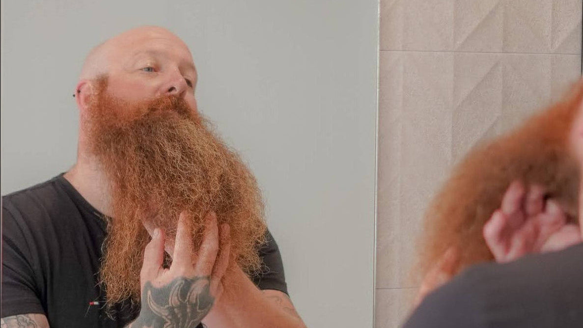 A man with a very large, full red beard and a shaved head stands in front of a bathroom mirror, touching and examining the beard. The back of his head and tattooed arms are visible, with his reflection shown in the arched mirror against a light tiled wall.