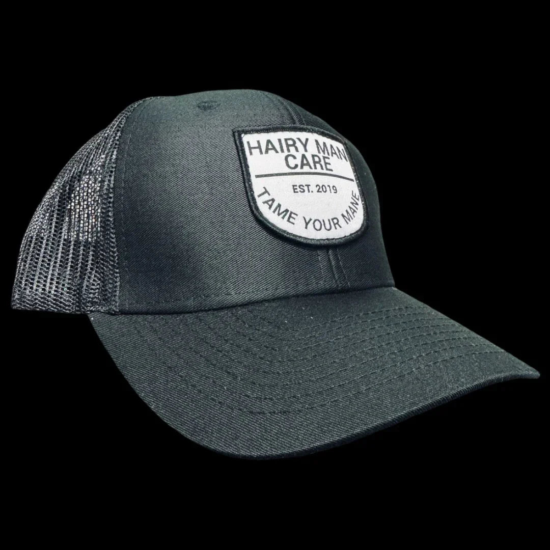 Dark gray trucker cap with Hairy Man Care patch, 'Tame Your Mane' slogan, mesh back, established 2019