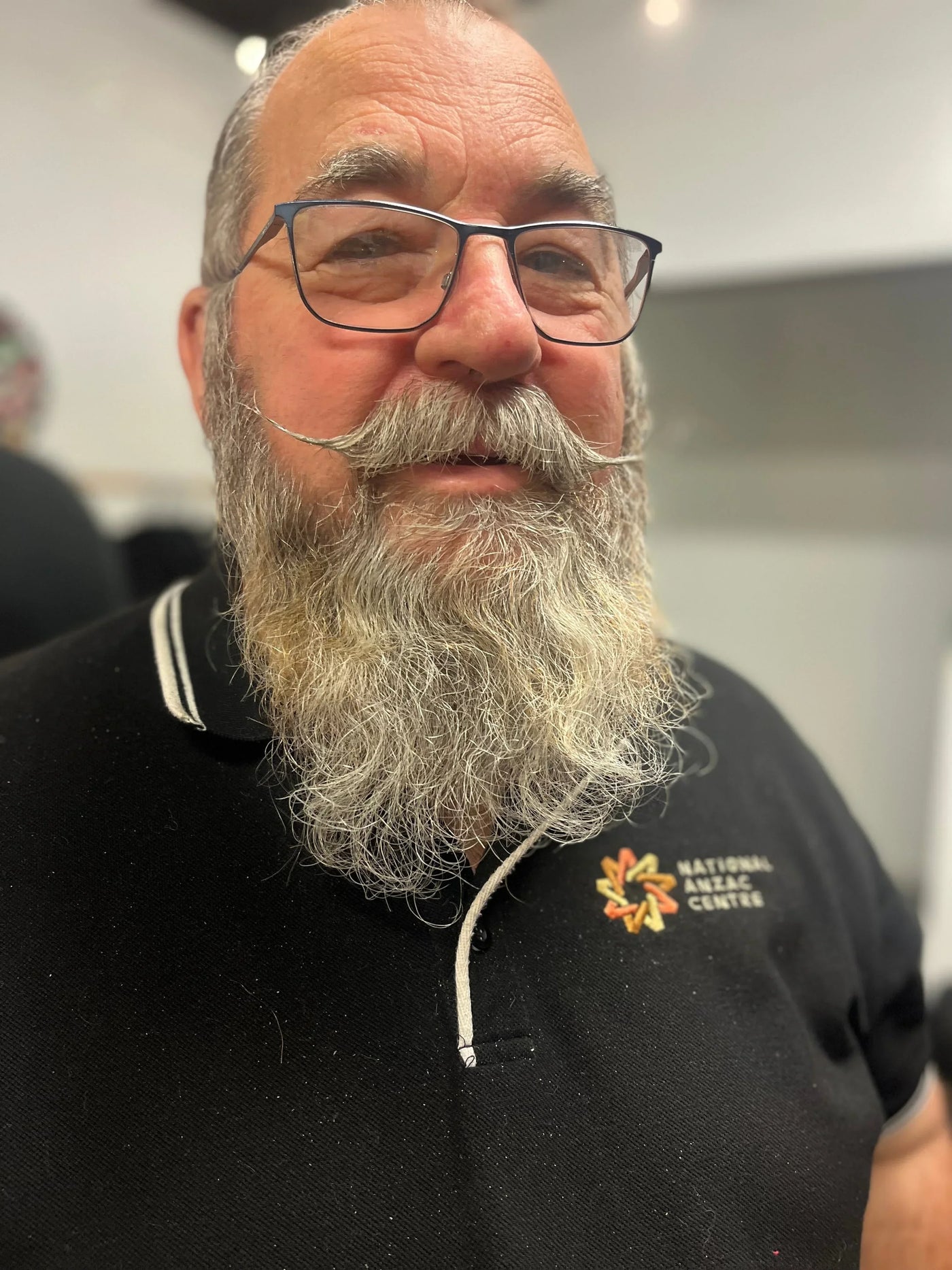 Man with full grey beard and glasses, wearing National Anzac Centre polo shirt